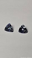 Pair of triangular, trillion cut tanzanite gemstones on a plain white background displaying their facets and color spectrum of purple and blue.