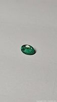 Close-up photo of a natural oval cut green emerald on plain light surface, showing color and internal characteristics.
