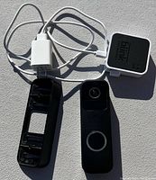 Photo of Blink doorbell camera unit, its black mounting bracket, white USB charging cable, white power adapter, and Blink Sync Module 2 unit.