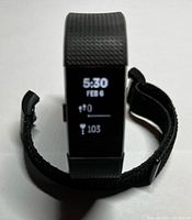 Fitbit Charge 2 fitness tracker powered on showing time, date, steps, and heart rate on display. Black ridged wristband attached, extra fabric band beside it.