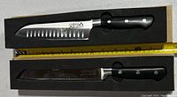 Two knives in foam cutouts with measuring tape for scale, brand 'cooking pleasures' visible on blade and handle. Santoku style blade with scalloped edge and black handle with three rivets.