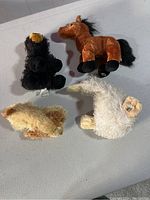 Four assorted stuffed animals arranged on a flat surface: black bear, brown horse, yellowish plush animal, fluffy white sheep.