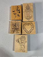 Front view of the top faces of the 5 wooden block rubber stamps showing detailed black outlines of their designs: leafy branch, ornate shield, flowers, heart with ruffle, and cherub angel.