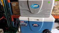 Photo showing two stacked coolers; the bottom is a blue and white Coleman Wheeled Xtreme 5 cooler, the top is a grey Xtreme cooler without wheels.