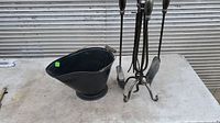Photo showing the black metal ash bucket and the fireplace tool stand with four tools on a concrete surface.