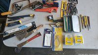 Overview of assorted hand tools and hardware on a white table surface including saws, drills, knives, pencils, and packaging for blades and bits.