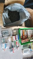 Cardboard box with plastic liner filled with a large number of metal nails and small packaged boxes with fasteners arranged on a surface.
