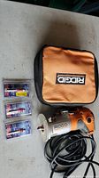 Photo showing RIDGID palm router, orange black carrying case, and three Freud router bits in packaging laid on a gray table