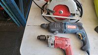 Top-down view of Milwaukee circular saw, Ryobi hammer drill and Milwaukee drill on a table showing all three tools with cords and drill bits attached.