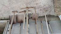 Full lot overview showing sledgehammer, picks, shovels, rakes, and pitch forks laid out on ground and metal shelf