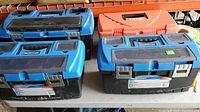 Four plastic toolboxes, three blue and black Mastercraft and one orange, arranged in two rows on a shelf.