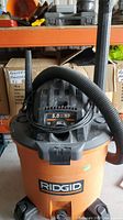 Front view of Rigid wet/dry vacuum showing black hose and orange body with Rigid logo.