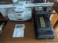 Antique cast iron adding machine with cream-colored keys and manual lever next to white Aiwa boombox with stereo speakers, remote control, and instruction manual.