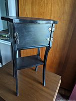 Front and angled view of black antique humidor cabinet showing the upper cabinet with a closed hinged door and lower open shelf. Original metal hinges and latch hardware visible.
