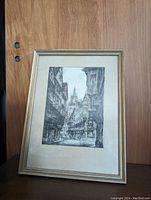 Framed monochrome etching print displayed against a wooden background. The artwork shows a street view with buildings and detailed architectural elements in black ink on handmade paper with a mat and frame.