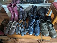 Lot arranged on bench showing front view of nine pairs of shoes and boots including purple rain boots, black boots, sandals, and orthopedics