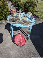 Items arranged on and around a light blue outdoor table including birdhouse, metal plaques, angel planter, red hanging planter, and metal stakes.