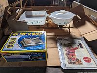 Photo showing two Glasbake casseroles with snowflake pattern, metal burner rack, unopened boxed Top-O-Matic cigarette machine, and vintage Santa advertisement.
