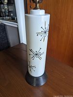 Mid century style Spudnik lamp showing white base with black and gold starburst hand painted motifs on cylindrical lamp body.