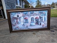 Front side showing a detailed scenic picture of a European-style café with arched windows, red awnings, outdoor seating, and floral decorations.