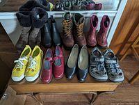Nine pairs of assorted boots and shoes displayed on a wooden table including Uggs, Wolverine boots, Bogs toddler rain boots, yellow Cariuma sneakers, and Teva sandals.