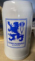 Front side of the ceramic beer stein showing the blue Lowenbrau Munich lion logo.