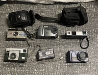 Six cameras and two cases arranged on a grey quilted surface including Kodak and Samsung models