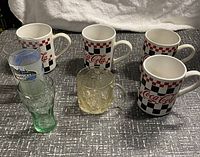 Overview of four Coca-Cola ceramic mugs with checkerboard pattern and three clear glass Coca-Cola drinking glasses