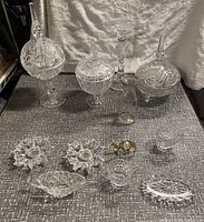 Full lot of crystal and glass candy dishes and decorative items on display on a textured surface with a silver-colored fabric backdrop.