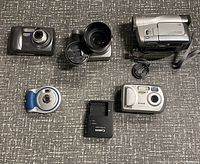 Top down photo showing all items in the lot including five cameras and one Canon battery charger on a carpeted surface