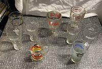 Photo showing assortment of 10 vintage collectible glasses including Coca-Cola branded glasses, Garfield character mug, and Miller Lite shot glass on a gray patterned surface.