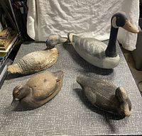 Lot of four carved wooden bird decoys on tabletop: Canada goose, and three ducks in various wear and paint conditions.