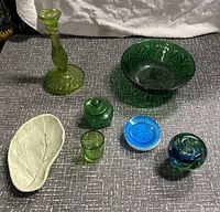 Overview photo of the lot showing green glass candlestick, large green bowl, small green jar with lid, small green tumbler, blue glass dish with white rim, glass apple paperweight, and white leaf-shaped pottery dish.