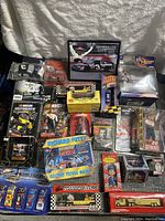 NASCAR collectibles including diecast cars, figures, VHS tapes, trivia game, and memorabilia are shown arranged on a gray surface. Items include racing trivia game box, collectible figures, multiple diecast cars in various packaging including Tony Stewart and Ernie Irvan editions.