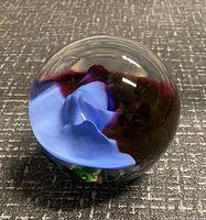 Photo of a clear glass paperweight with an internal blue swirl and dark red shapes inside, placed on a textured dark surface.