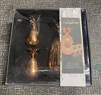 Top view of the pineapple bar set showing copper tone pineapple tumbler and two shot glasses in black molded plastic packaging with a label describing contents.