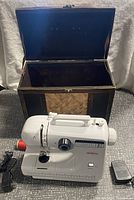 Front view of white Sunbeam sewing machine with brown woven vintage carrying case open behind it. Machine is plugged into a power cord, foot pedal to the right.