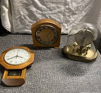 Three vintage clocks on a gray fabric surface with white cloth background: a wooden wall clock with Roman numerals, a square wooden mantel clock with Arabic numerals, and an anniversary clock under a glass dome.