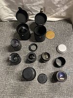 Top-down view of seven vintage camera lenses, two black padded cylindrical lens cases, and small caps on gray woven surface.