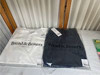 Two sealed clothing items in plastic packaging, a white shirt and a dark-colored pair of shorts with Bread & Boxers branding.