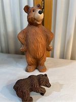 Two bear figurines on a white cloth surface with curtains and a door in the background. Larger bear stands upright while smaller bear is positioned in front of it on all fours.