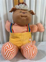 Full frontal view of the vintage Cabbage Patch Kids plush doll, seated, showing striped arms and legs, yellow overalls with horse patch, and yarn hair.
