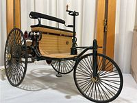Side view of the diecast 1886 Mercedes-Benz model showing the wooden floorboard, black metal frame, spoked wheels, and bench seat.