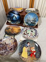 Photo of various decorative plates including nature-themed scenes with ships and wildlife, and Disney character scenes Snow White and others. Plates are displayed on a white tablecloth, some in wooden stands.