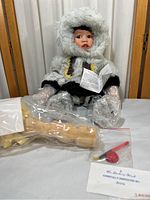 Front view of porcelain Indigenous doll dressed in winter parka, wrapped in protective plastic at hands and feet, with wooden carved accessory and small parts in plastic bags in front.