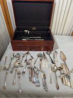 Wooden silverware case open with multiple silver plated flatware pieces laid in front including spoons, forks, and knives, showing general condition and content of lot.