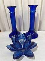 Front view of two cobalt blue ribbed glass candlesticks and one faceted blue glass lotus flower decorative piece arranged on white fabric.