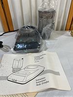 Ultrasonic jewelry cleaner SM 360 with plastic basket insert wrapped in clear plastic, power cord visible, and instruction manual in front.