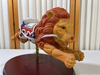 Front angled view of leaping lion figurine showing detailed face, mane, harness and saddle, on round wooden base.