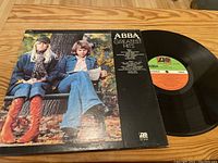 Front cover of ABBA Greatest Hits vinyl record with track listing and image of two band members seated outdoors.